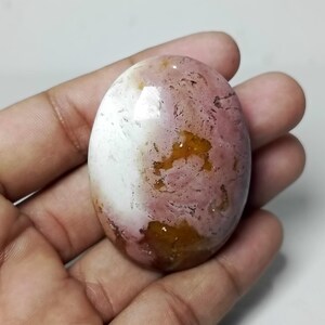 May include: Oval-shaped gemstone with a mix of colors. The stone features a blend of white, pink, and golden-brown hues. The polished surface reflects light, highlighting the natural patterns and textures within the stone. The gemstone is held in a hand.