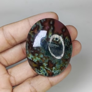 May include: Oval-shaped gemstone with a polished surface. The stone features a mix of colors, including deep red, green, and brown, with a speckled pattern. The gemstone is held in a hand, showcasing its size and shape.