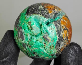 Puede incluir: Una gema esférica con una mezcla de colores, incluyendo verde vibrante, naranja, negro y marrón. La piedra tiene una superficie rugosa y texturizada y parece ser un espécimen mineral natural. La gema se sostiene en una mano enguantada.