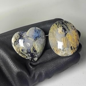 May include: Two polished gemstones: one heart-shaped and one oval. The heart-shaped stone has a mix of blue, white, and black patterns. The oval stone features yellow, black, and white patterns. Both stones are smooth and glossy.