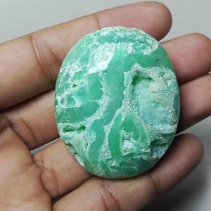 May include: Oval-shaped, light green gemstone with a rough, textured surface. The stone has areas of lighter green and white, creating a marbled effect. The gemstone is held in a hand, showcasing its natural beauty.
