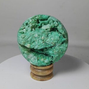 May include: A spherical, rough-textured, green mineral specimen, possibly a geode, sits atop a small, wooden, tiered stand. The mineral's surface is a mix of vibrant green and lighter, crystalline areas, creating a visually interesting contrast.