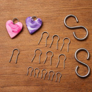 Peut inclure: Divers éléments de bricolage sur une surface en bois. L'image présente deux pendentifs en forme de cœur avec des motifs roses et violets, plusieurs crochets en S argentés et de nombreuses attaches en forme d'épingle à cheveux argentées.
