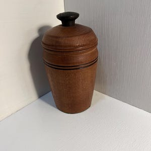 May include: A wooden container with a lid. The container is a warm brown color with a black knob on top. Two black lines encircle the body of the container. The container is sitting on a white surface.