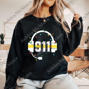 May include: Black sweatshirt featuring a white headset design with the number "911" in the center. The headset is decorated with colorful string lights. The design includes a microphone. The sweatshirt is a crew neck.
