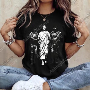 K&ouml;nnte beinhalten: Schwarzes T-Shirt mit einer Grafik von Jesus, flankiert von zwei Figuren in taktischer Ausr&uuml;stung, jeweils mit "ICE" auf ihrer Uniform. Das Bild ist in Schwarzwei&szlig;.