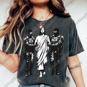 K&ouml;nnte beinhalten: Dunkelgraues T-Shirt mit einer Grafik von Jesus, flankiert von zwei Figuren in taktischer Ausr&uuml;stung, jeweils mit "ICE" auf ihren Westen. Die zentrale Figur ist wei&szlig;, im Kontrast zu den schwarzen Figuren.