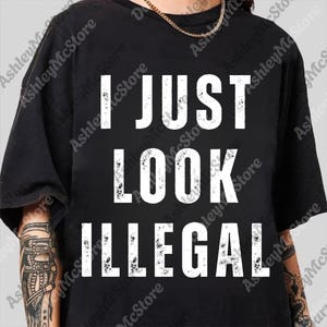 Peut inclure: T-shirt noir avec le texte blanc "I JUST LOOK ILLEGAL". Le texte est en gras, en police sans-serif. Le t-shirt a des manches courtes et un col rond.