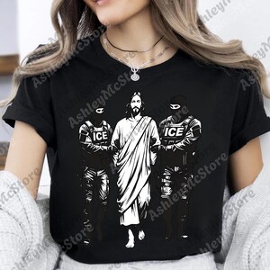 K&ouml;nnte beinhalten: Schwarzes T-Shirt mit einer wei&szlig;en Grafik, die Jesus zwischen zwei Figuren in taktischer Ausr&uuml;stung zeigt, jede mit "ICE" auf ihrer Uniform. Ein auff&auml;lliges, grafisches Design.