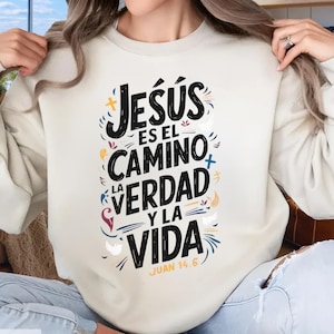 May include: Cream-colored sweatshirt with the text "Jesús es el Camino, la Verdad y la Vida" in black, with colorful accents. The text is surrounded by decorative crosses, leaves, and other small designs. The text "Juan 14.6" is at the bottom.