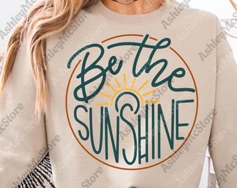 May include: A beige sweatshirt with the words "Be the Sunshine" in a circular design. The text is in teal script with a yellow sun graphic in the center. The sweatshirt is a neutral color.