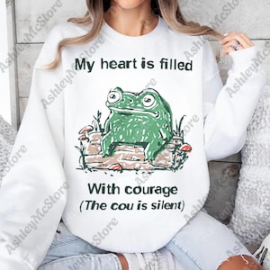 Op de afbeelding: Witte sweatshirt met een groene kikkerillustratie op een boomstam, omringd door gras en paddenstoelen. De tekst "My heart is filled with courage (The cou is silent)" staat erboven en eronder.