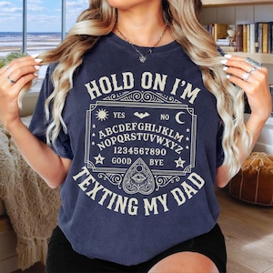 May include: A dark blue t-shirt featuring a cream-colored graphic of a spirit board with the alphabet, numbers, and the text 'HOLD ON I'M TEXTING MY DAD' arranged around a central planchette icon with an eye design and decorative border elements.