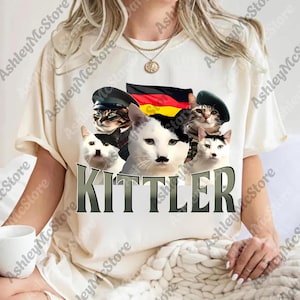 May include: White t-shirt featuring a graphic of several cats wearing hats and a German flag. One cat has a mustache. The word "KITTLER" is printed in large letters below the cats.