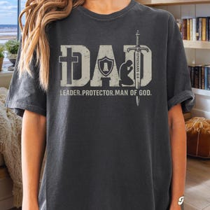 May include: Dark grey t-shirt with the word "DAD" in a distressed, light grey font, featuring a cross, shield, and kneeling figure. Below, the text reads "LEADER. PROTECTOR. MAN OF GOD." A sword graphic is on the right side.