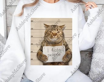 May include: White sweatshirt featuring a humorous graphic of a cat posing for a mugshot, holding a sign that reads "I hate people." The cat has brown and tan fur and is set against a height chart background.