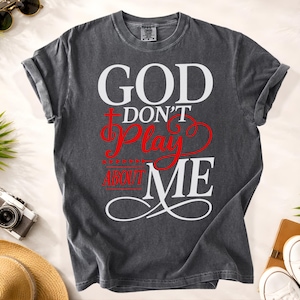 May include: Dark gray t-shirt with the text "GOD DON'T PLAY ABOUT ME" in white and red lettering. The shirt has a crew neck and short sleeves, and is made of a soft, comfortable material.