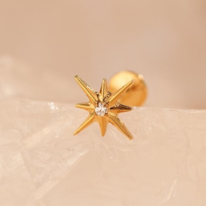May include: A gold stud earring with a starburst design and a small, clear gemstone. The earring has a post and backing. The jewellery is set against a soft, neutral background.
