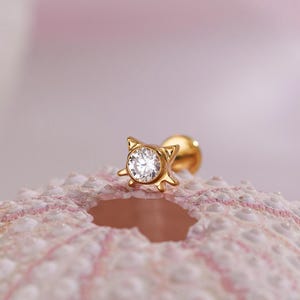 Op de afbeelding: Een gouden oorbel met een heldere, ronde edelsteen in het midden. De oorbel heeft een katvormig ontwerp. Het wordt getoond op een roze en wit getextureerd oppervlak tegen een zachte roze achtergrond.