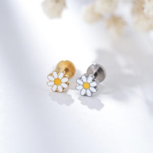 Puede incluir: Dos pendientes con forma de margarita. Uno es dorado, el otro plateado. Ambos tienen pétalos blancos y un centro amarillo. Los pendientes están diseñados para un piercing en el labio o en la oreja.
