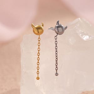 Puede incluir: Un par de pendientes desiguales. Uno es dorado con un diseño de cuernos de diablo y una cadena. El otro es plateado con un diseño de pájaro y una cadena. Ambos pendientes están sobre una superficie cristalina blanca.