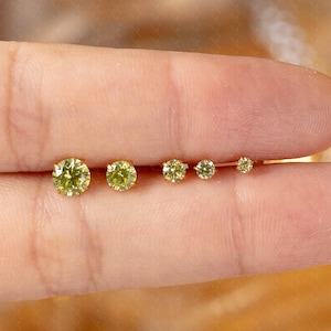 Könnte beinhalten: Eine Sammlung von fünf gelb-grünen Edelstein-Ohrsteckern in verschiedenen Größen. Die Ohrringe sind in Gold gefasst und vor einem verschwommenen, warmtonigen Hintergrund dargestellt. Die Edelsteine sind rund geschliffen.