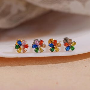 May include: Four flower-shaped stud earrings with colorful gemstone petals. The earrings feature a gold or silver setting and a central clear stone. The gemstones are arranged in a floral pattern, showcasing a variety of colors.
