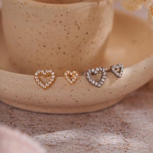 Puede incluir: Cuatro pendientes en forma de corazón. Dos son dorados y dos plateados. Cada pendiente está incrustado con pequeños cristales transparentes. Los pendientes se exhiben en un plato de cerámica beige.