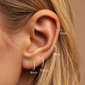 May include: A close-up of an ear adorned with several small, gold hoop earrings. The earrings are encrusted with small, sparkling stones. Sizes are labeled as 6mm, 8mm, and 10mm. The ear is set against a soft, neutral background.