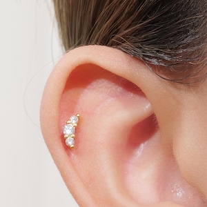 Puede incluir: Un piercing dorado con tres gemas redondas y transparentes dispuestas en una línea curva. La joya está colocada en la oreja, destacando las piedras brillantes y el diseño delicado. Este pendiente es una pieza de joyería corporal.