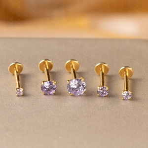 Può includere: Cinque orecchini a bottone dorati con retro piatto e perno. Ogni orecchino presenta una gemma rotonda di colore viola chiaro. Gli orecchini sono disposti in fila, mostrando variazioni di dimensioni.