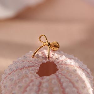 May include: A gold-colored bow-shaped earring stud. The earring is set on a light pink and white textured surface. The bow design is delicate and detailed, with a small post for insertion.