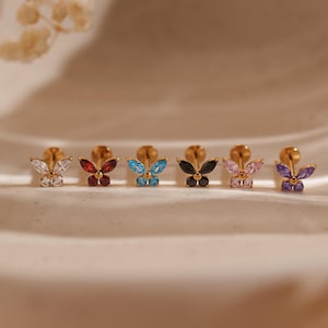 Könnte beinhalten: Sechs goldfarbene Ohrstecker in Schmetterlingsform mit farbigen Edelsteinflügeln in Weiß, Rot, Blau, Schwarz, Rosa und Lila. Die Ohrringe sind auf einer weißen Oberfläche ausgestellt.