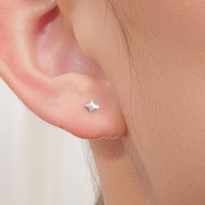 May include: A close-up of a person's ear with a small, silver star-shaped stud earring. The earring is a four-pointed star with a slightly textured surface, reflecting light. The skin around the ear is smooth and pale.