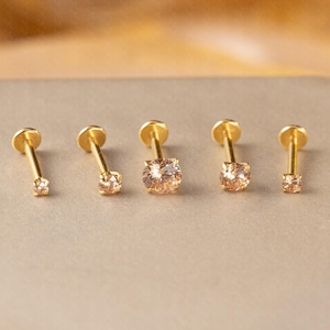 Puede incluir: Cinco pendientes dorados con parte trasera plana y gemas redondas de color naranja claro de diferentes tamaños. Los pendientes están dispuestos en fila, mostrando sus diferentes dimensiones y diseños. El fondo es neutro.