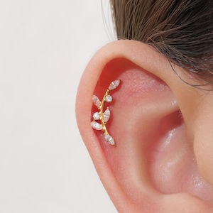 May include: A gold-colored ear piercing featuring a delicate leaf design. The piercing is adorned with clear, sparkling gemstones, arranged to mimic leaves and small round accents. The jewelry is set against the skin of an ear.