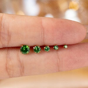 May include: Five gold stud earrings with green gemstones of varying sizes. The earrings are displayed in a line, with the gemstones getting smaller. The round, faceted gems sparkle. The gold settings provide a classic look. The earrings are a jewellery item.