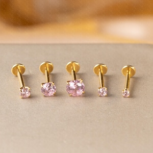 May include: Five gold-colored stud earrings with pink gemstones. The earrings have a flat disc backing and a post. The gemstones are round or square-cut. The earrings are arranged in a row on a neutral surface.