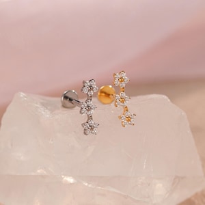May include: Two flower-shaped earrings. One is silver-toned, the other gold-toned. Each earring features three small, sparkling flower clusters. The earrings are displayed on a white, textured surface against a soft pink background.