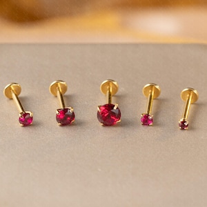 May include: Five gold-coloured stud earrings with vibrant red gemstones. The earrings are arranged in a row, showcasing variations in gemstone size and design. The studs are set against a neutral background.