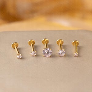 May include: Five gold-coloured stud earrings with clear, round gemstones. The earrings are arranged in a row, showcasing variations in gemstone size. The studs have a flat back and a post for piercing.