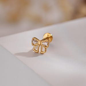 Könnte beinhalten: Goldfarbener Ohrring in Schleifenform mit zwei klaren, rechteckigen Edelsteinen. Der Ohrring hat einen Stecker und einen runden Verschluss. Das Schleifendesign ist offen.