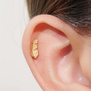 May include: A gold-colored feather-shaped earring stud is shown in a close-up view. The earring is small and detailed, with a textured surface that mimics the appearance of a feather. The earring is placed on the ear of a person.
