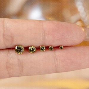 May include: A set of five gold-colored stud earrings with olive-green gemstones of varying sizes. The earrings are arranged in a row, showcasing the graduated sizes of the round-cut stones. The earrings are displayed on a person's skin.