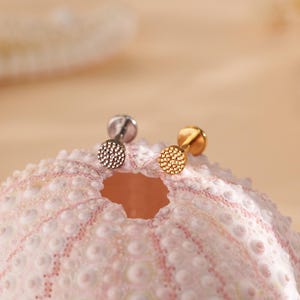 May include: Two stud earrings, one silver and one gold, each with a textured, circular design. The earrings are displayed on a pink and white textured surface, with a blurred background.