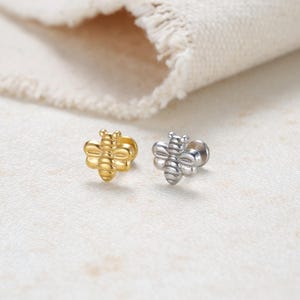 Puede incluir: Dos pendientes de botón con forma de abeja. Uno es dorado y el otro plateado. Los pendientes son pequeños y detallados, con alas y segmentos corporales visibles. El fondo es una superficie clara y texturizada.