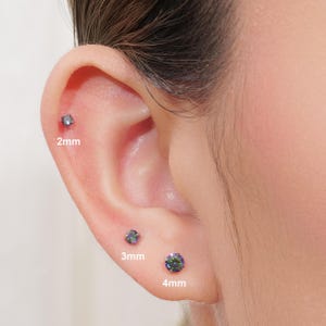 May include: Close-up of an ear with three iridescent stud earrings. The studs are labeled 2mm, 3mm, and 4mm. The earrings have a shimmering, multi-colored appearance, reflecting various hues.