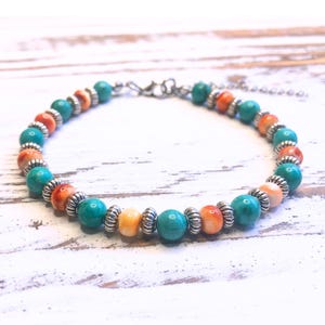 May include: A beaded bracelet featuring turquoise, orange, and yellow beads, separated by silver-colored metal spacers. The bracelet is strung on a silver chain and rests on a white wooden surface.