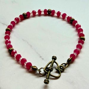 May include: A beaded bracelet featuring faceted red beads and clear spacer beads. The bracelet includes antique brass-colored accents and a heart-shaped toggle clasp. The jewelry is displayed on a white marble surface.