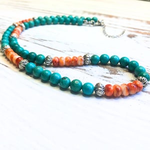 May include: A turquoise and orange beaded necklace with silver accents. The necklace features round turquoise beads interspersed with orange beads and decorative silver beads. The necklace is displayed on a white wooden surface.